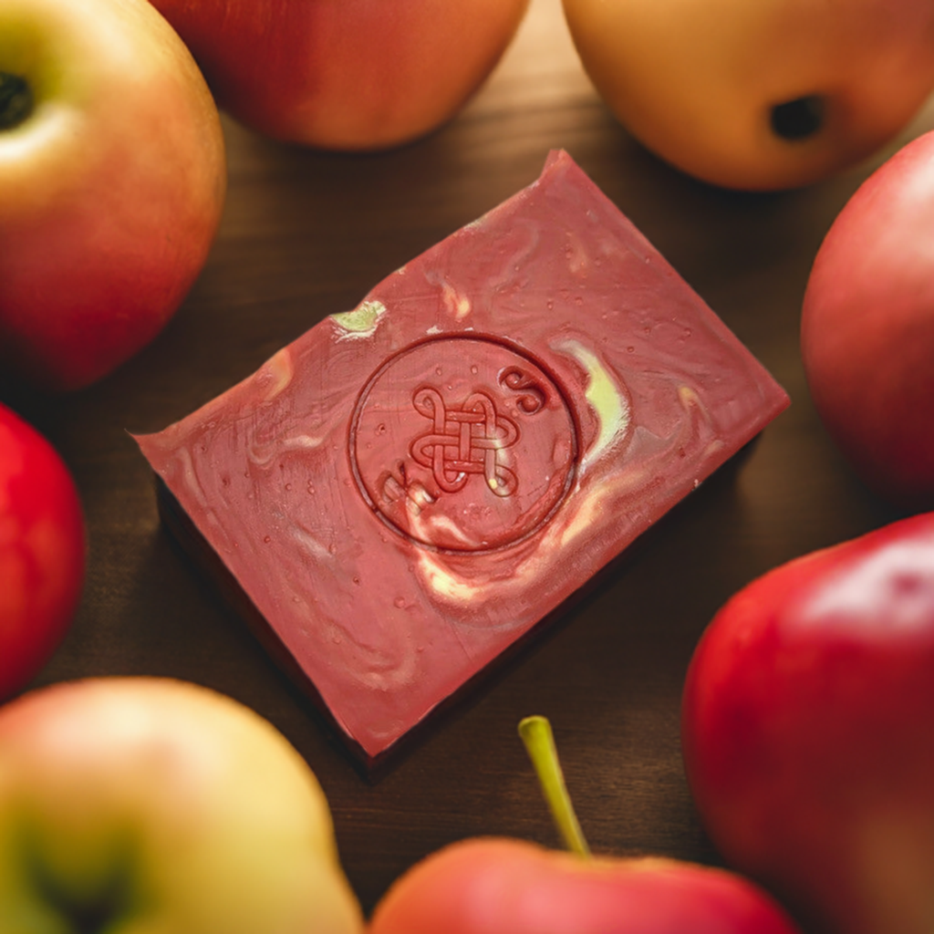 A bar of soap with a swirled red and yellow pattern, featuring the logo of the brand on top, placed on a wooden surface surrounded by various apples.